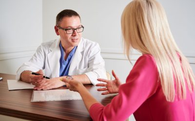 Male surgeon in white lab coat sitting at the table with papers and talking with female patient in office, discussing plastic surgery. Concept of medical appointment and cosmetic surgery.