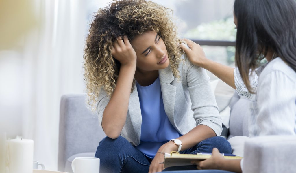 A young woman sits on a couch with her unrecognizable therapist.  She puts her head in her hand as she looks out the window with a sad expression.  Her therapist puts a hand on her shoulder.